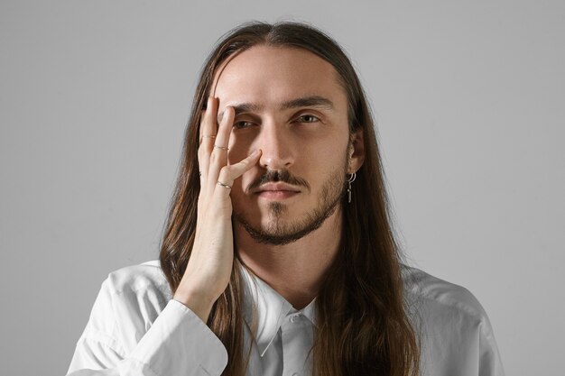 Human facial expressions. Isolated shot of handsome unshaven young Caucasian male with long hair posing, having serious look, holding hand on his face, wearing stylish shirt and accessories