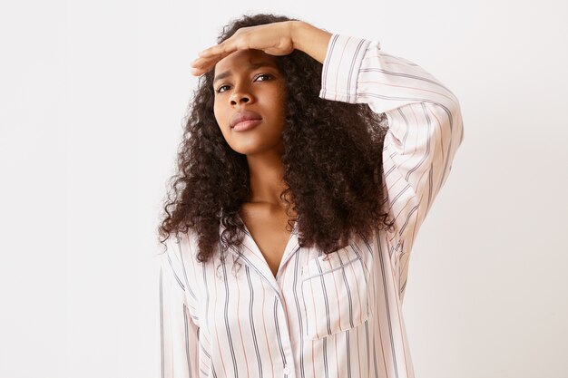 Human facial expressions, reaction and emotions. Attractive young African woman in stylish pajamas holding palm at her forehead, looking into distance, trying to see clearly, having concentrated look