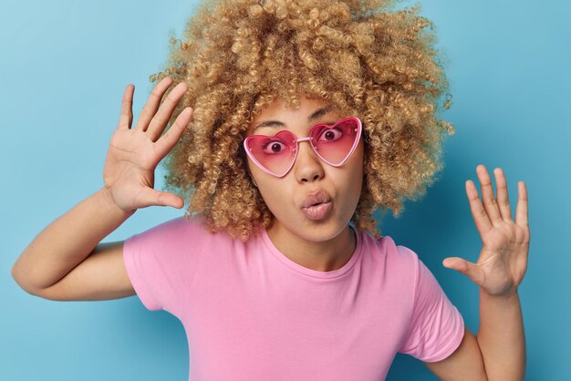 Human facial expressions and reactions concept Curly haired woman keeps lips folded palms raised looks surprisingly at camera wears trendy pink sunglasses and t shirt isolated over blue background