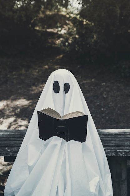 Human in ghost suit sitting on bench and reading book