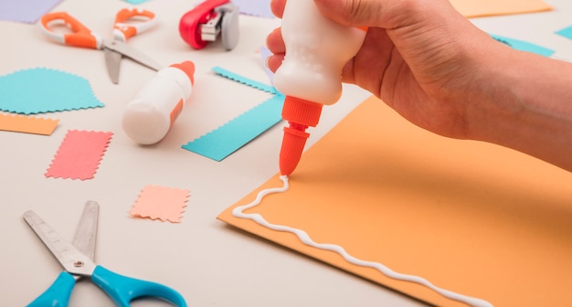 Human hand applying white glue on orange paper with scissor and stapler