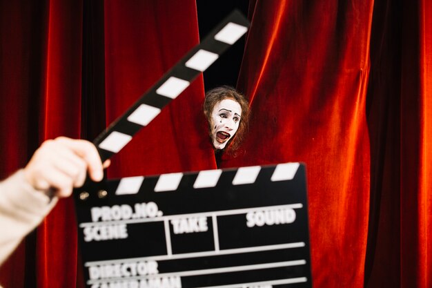 Human hand holding clapperboard in front of male mime artist performing behind red curtain