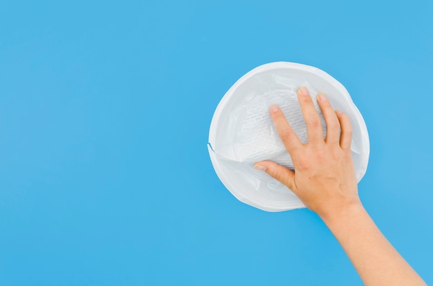 Human hand holding crumpled plastic plate on blue surface