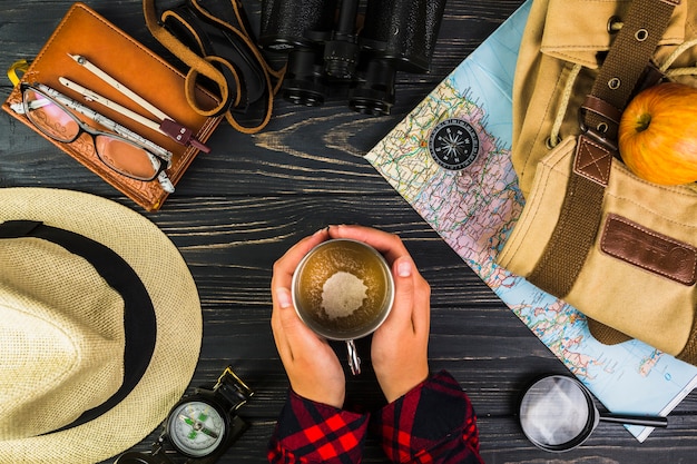 Human hand holding cup of coffee surrounded with various travelling equipments