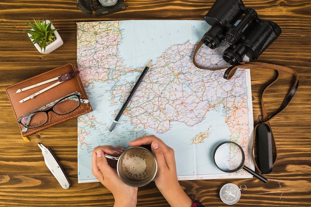 Human hand holding cup of coffee with travelling equipments on desk