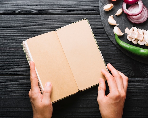 Free photo human hand holding diary near chili pepper; garlic cloves; onions and mushrooms on black wooden surface