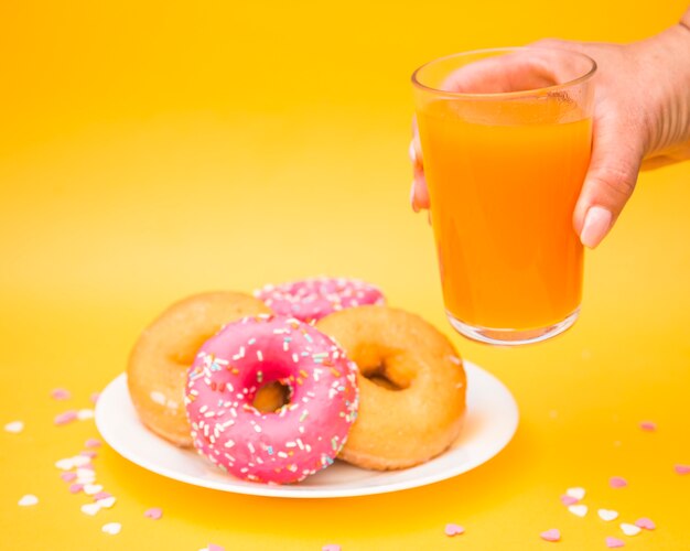 Human hand holding glass of juice near donuts on plate