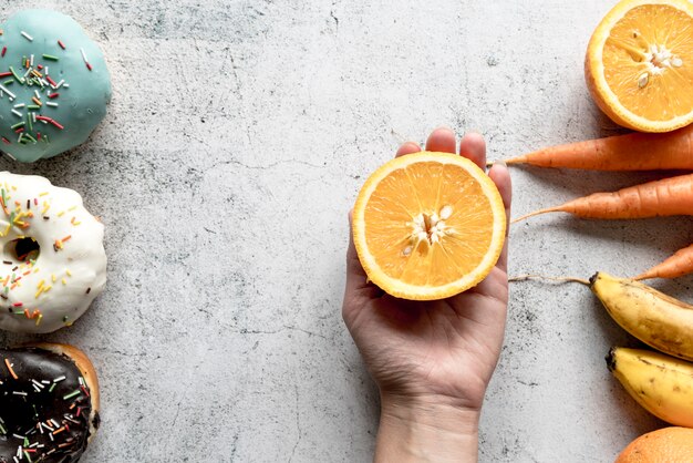 Human hand holding halved orange fruit near donuts; carrots and banana