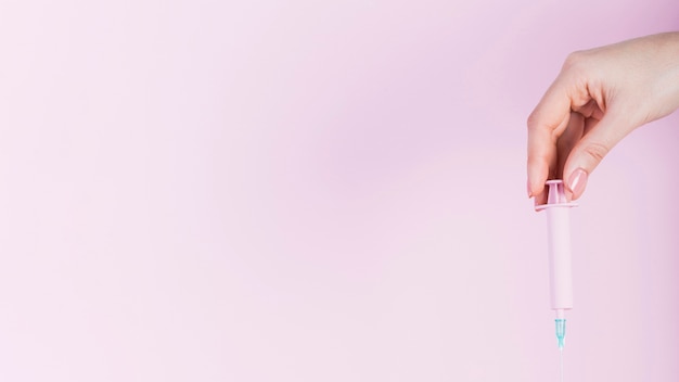 Human hand holding plastic syringe over pink backdrop