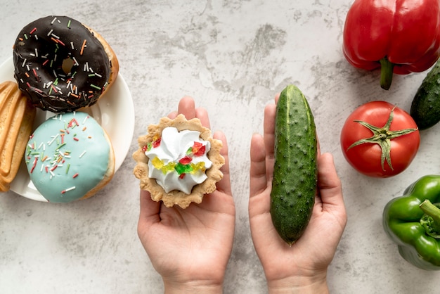 Human hand holding tart cake and cucumber near vegetables and sweet food over background