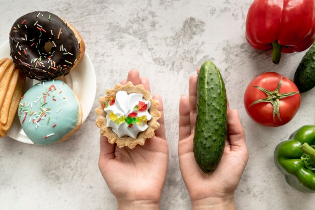 Human hand holding tart cake and cucumber near vegetables and sweet food over background