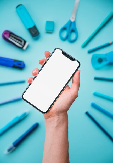 Human hand holding white screen cellphone over stationery arranged in circular shape on blue backdrop