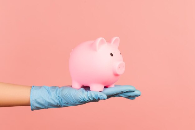 Человеческая рука в синих хирургических перчатках держит копилку свиньи. экономия, финансовая или банковская концепция.
