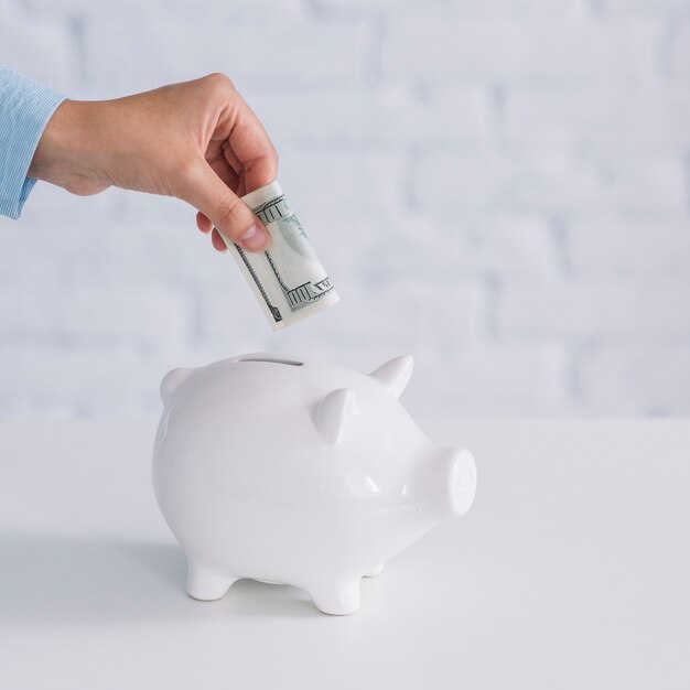 Human hand inserting banknote in white piggybank on desk