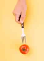 Free photo human hand inserting fork in red tomato on yellow backdrop