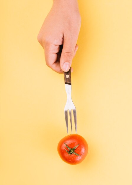 Human hand inserting fork in red tomato on yellow backdrop