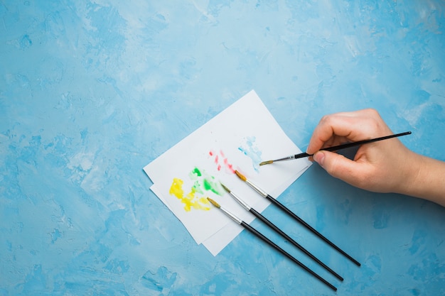 Human hand painting on white sheet with paint brush over blue pastel backdrop