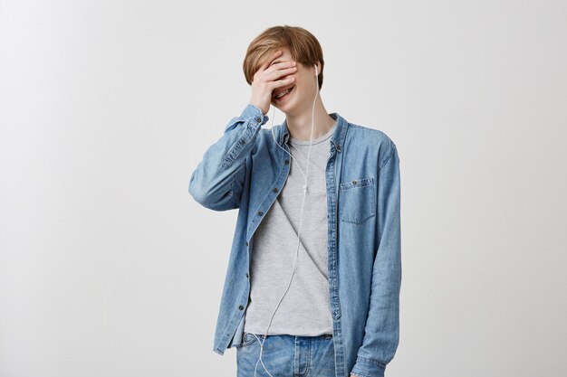 Human, leisure and modern technology concept. Indoor shot of fair-haired young male in denim shirt, listens songs with headphones, doesn`t hear anything around, hiding his face behind his palm.