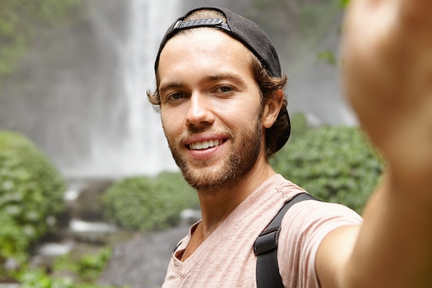 Human, nature and tourism. Handsome young traveler in snapback smiling joyfully while taking selfie, posing by gorgeous waterfall in rainforest