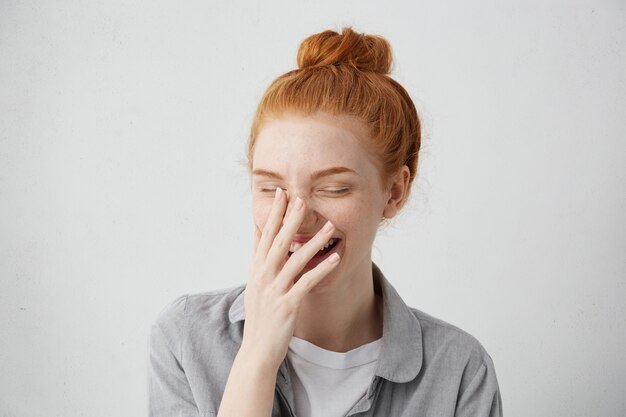 인간의 긍정적 인 감정과 감정. 손으로 입을 벌리고 눈을 감고 그녀의 마음의 바닥에서 웃고있는 머리카락 매듭을 가진 귀여운 소심한 매력적인 젊은 유럽 여성의 얼굴 사진