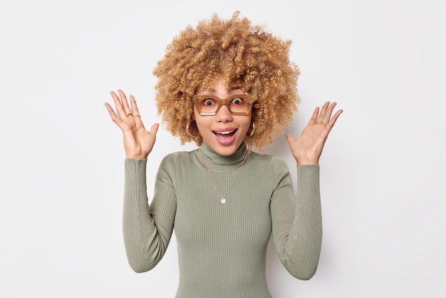 Human reactions concept. Emotional curly haired woman looks with great wonder at camera keeps palms raised feels fascinated reacts on unexpected offer wears spectacles casual jumper poses indoor