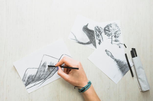 Human's hand sketching on paper with charcoal stick near beautiful hand drawn drawing