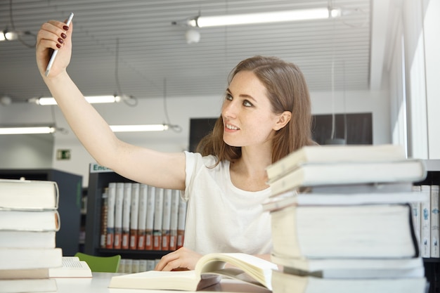 Human and technology. People and education. Indoor portrait of Caucasian teenage woman at library trying to take a selfie, surrounded by books and manuals