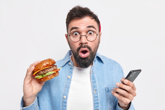 Hungry bearded adult man eats delicious burger holds mobile phone finds out shocking news wears round spectacles denim shirt 