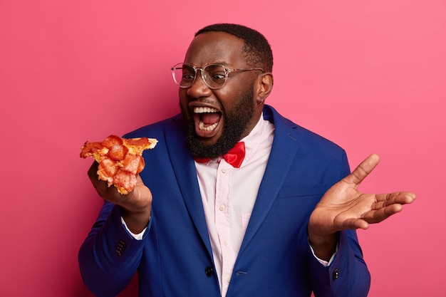 배고픈 흑인은 매우 큰 피자 조각을 물고, 식욕이 있으며, 공식적인 옷을 입고 안경을 쓰고 분홍색 공간에 포즈를 취합니다.