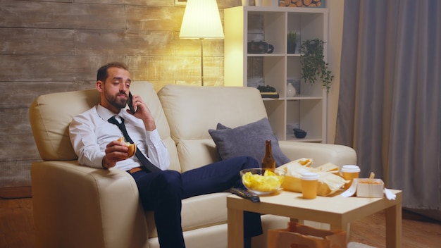 Hungry businessman having a conversation on his mobile phone while eating a burger. Beer on the table.