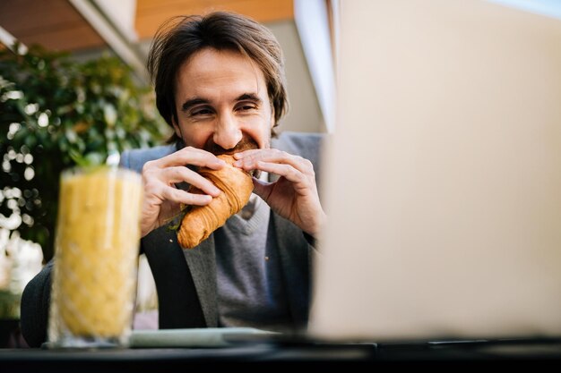 Голодный бизнесмен ест круассан, читая электронную почту на ноутбуке в кафе