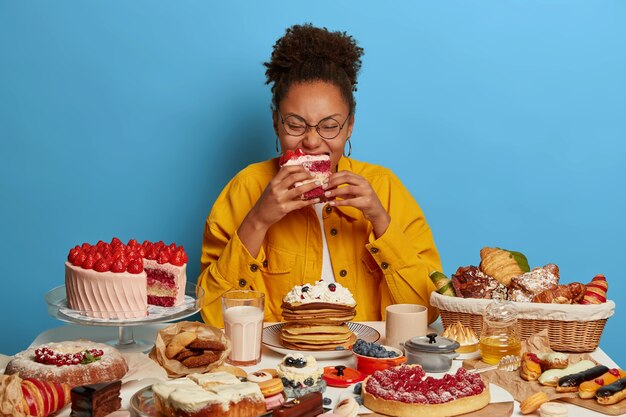 Hungry curly woman eats with appetite creamy strawberry cake, has addiction to sugar, comes on brithday, tastes various desserts