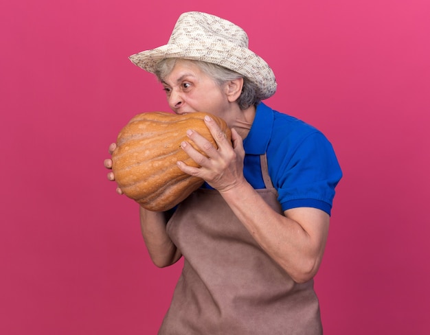 Hungry elderly female gardener wearing gardening hat holding and pretending to bite pumpkin
