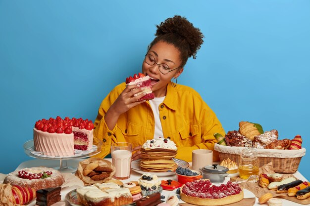 Hungry greedy Afro American girl bites big delicious piece of cake, poses at table with many yummy desserts, has sweet breakfast at home, unhealthy nutrition, isolated over blue wall