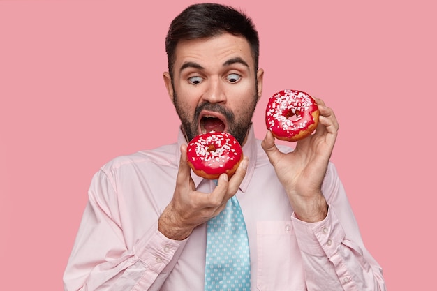 Free photo hungry handsome man bites red doughnut, wears formal shirt with tie, opens mouth widely