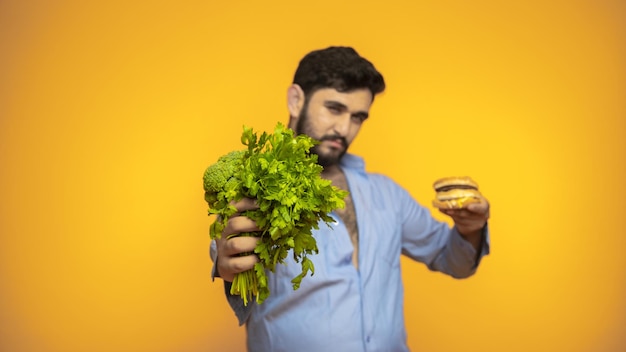 空腹のハンサムな男は、黄色の背景にハンバーガーを手に持っています