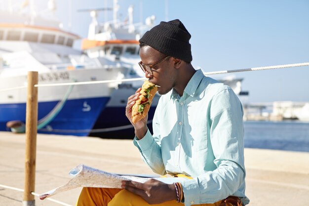 Hungry male tourist in trendy clothing and accessories eating a sandwich