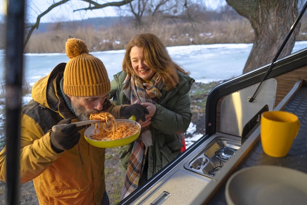 Hungry man eat lunch pasta from pan outdoors in wild standing at mini camper back kitchen during winter journey vacation Family spend time camping away in the wild together Family journey concept