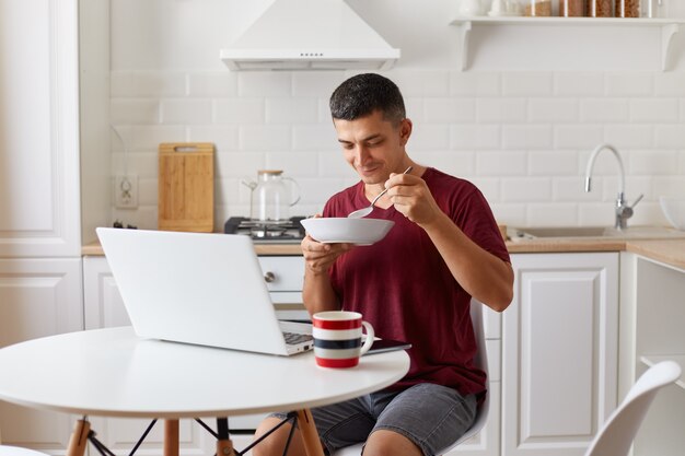 Hungry man freelancer sitting in kitchen at table in front of open laptop computer and eating soup, holding plate in hands, attractive guy wearing burgundy casual style t shirt.