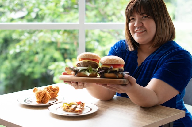Голодная женщина с избыточным весом, держащая гамбургер на деревянной тарелке жареный цыпленок и пицца на столе Premium Фотографии