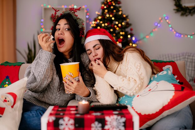 hungry pretty young girl with holly wreath eats popcorn and looks at her sleepy friend sitting on armchair and enjoying christmas time at home