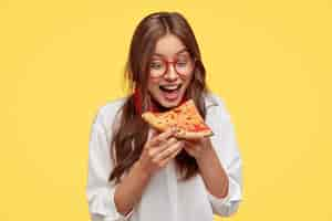 Free photo hungry student opens mouth widely while sees delicious slice of pizza, wants to eat, dressed in white shirt, models against yellow wall. positive woman with junk food. people and eating