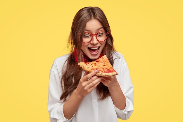 空腹の学生は、ピザのおいしいスライスを見て、食べたい、白いシャツを着て、黄色い壁にモデルを置いている間、口を大きく開きます。ジャンクフードとポジティブな女性。人と食事