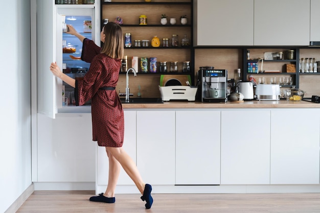 Голодная женщина ищет еду в холодильнике дома, но ее там мало Белая кухонная мебель, домашняя одежда, красный шелковый халат