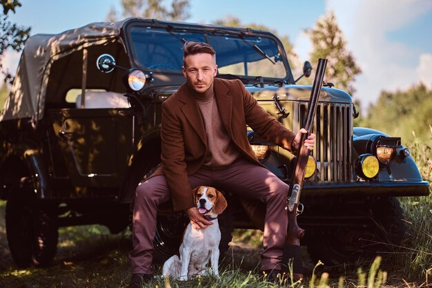 Hunter in elegant clothes holds a shotgun and sits together with his beagle dog while leaning against a retro military car in a forest.
