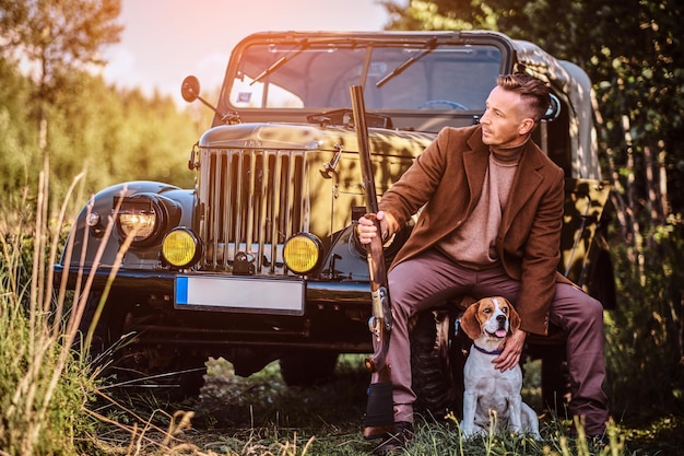 Hunter in elegant clothes holds a shotgun and sits together with his beagle dog while leaning against a retro military car in a forest.