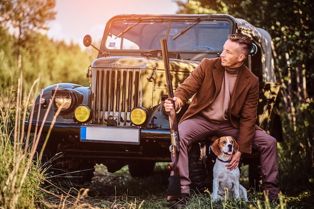 エレガントな服を着たハンターはショットガンを持って、森の中でレトロな軍用車に寄りかかっている間、ビーグル犬と一緒に座っています。