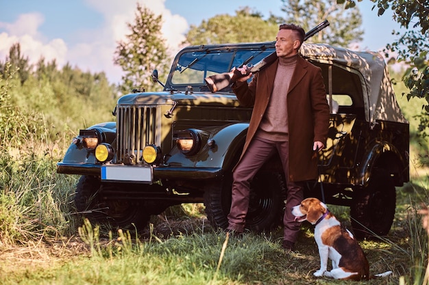Hunter in elegant clothes holds a shotgun and standing together with his beagle dog near a retro military car in the forest.