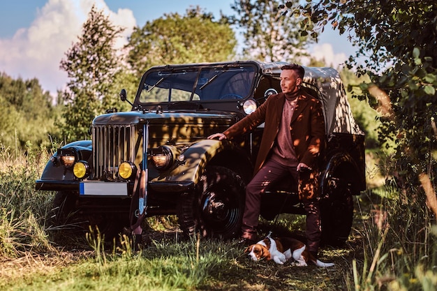 Hunter in elegant clothes standing together with his beagle dog near a retro military car in the forest.