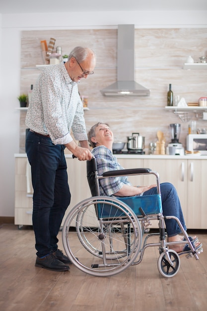 Husband looking at disabled senior woman in the kitchen. Disabled senior woman sitting in wheelchair in kitchen looking through window. Living with handicapped person. Husband helping wife with disabi